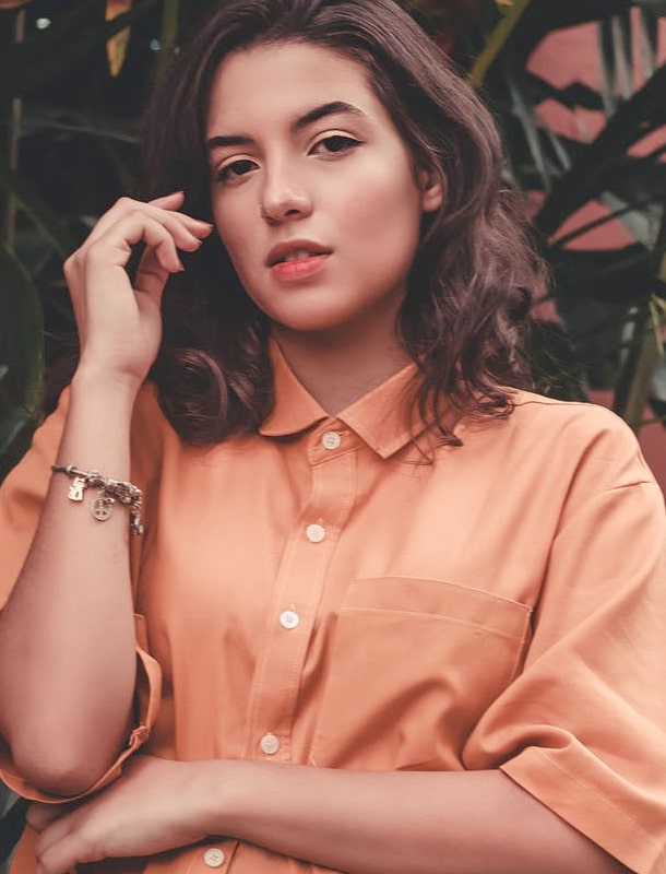 A young, black-haired women in a pink blouse stands in front of a tropical plant